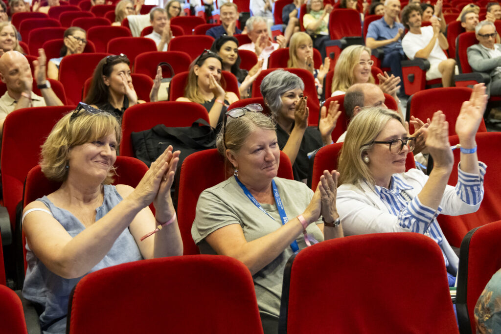 Audience in a theater clapping during an event; diverse group of adults in red seats.