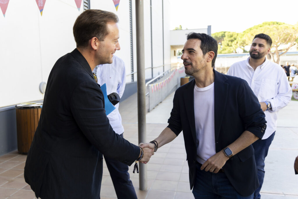 Two men in blazers shake hands outdoors while smiling; a third man watches in the background.