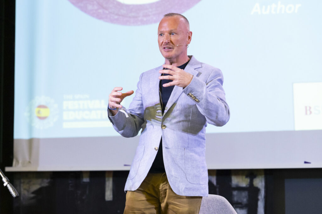 Male speaker on stage wearing a light blue blazer, gesturing as he talks into a headset microphone.
