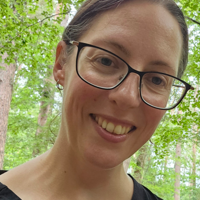 Smiling woman wearing black glasses outdoors among green trees.