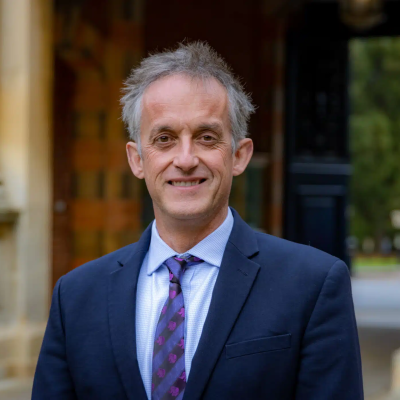 Smiling middle-aged man in a navy suit and purple patterned tie, outdoors in front of a stone building with columns.