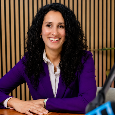 Smiling woman with curly black hair in a purple blazer at a desk, wood slat background, likely in a conference or interview setting